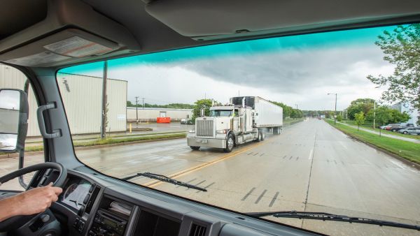 A view from inside a truck cab looking out at a two‑lane road on a cloudy day, with a white semi‑truck driving toward the camera and industrial buildings and greenery lining both sides of the street.